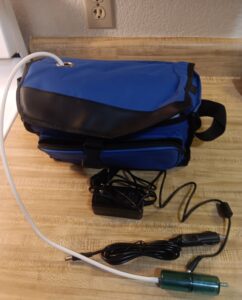 blue bag with a top pouch and a front zipper pouch. There is a cord coming out the top of the bag and there are also 2 cords on the table in front of the bag
