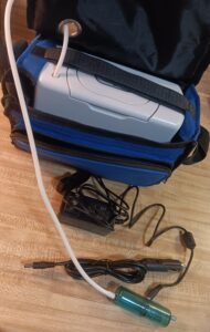 blue bag with top open showing a gray rectangular device inside with a small hose coming out of the top, and 2 cords in front of the bag