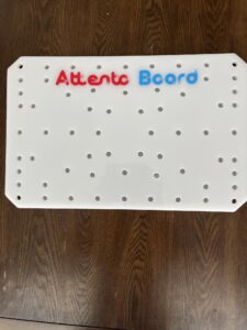 Rectangular white board with over 60 peg holes in it and text at the top of the board that says “Attento Board”
