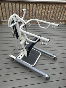 metal patient lift with u-shaped arm supports and handles, vertical metal bar that adjusts height, and a base with 4 wheels and a black foot platform