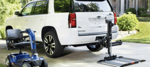 internet picture showing a wheelchair lift attached to the back of a vehicle, with the lift folded out and currently down on the ground. Electric wheelchair/scooter shown off to the side. Wheelchair/scooter would sit horizontal on the back of the vehicle