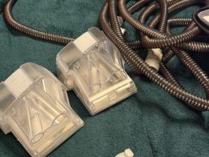 Two semi-transparent CPAP connectors sit on a blanket. The black tubes are lying next to the masks.