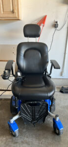 front view of an electric wheelchair with head rest, arm rests, and a foot rest. Wheelchair contains a blue base and a joy stick used for steering