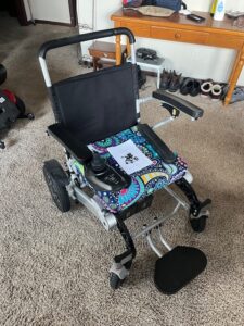 Small to medium-sized black electric wheelchair with floral seat cover. Gently used condition. An informational booklet is on the seat.