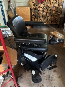 Side view of black electric wheelchair. Has four wheels, plus joystick and footrest. Seat has a smooth texture that may be leather. Pride and Go Chair logos are visible. Gently used condition.