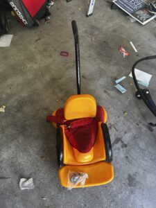 small orange seat with seatbelt and footrest. Seat contains 2 wheels and sits low to the ground. There is one handle bar on the back to push