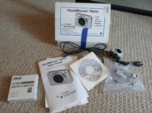 The following items are laid out on the floor: HeadMouse Nano with cord, user guide, software disc for SofType On-Screen keyboard, USB Wi-Fi adapter, mounting bracket, and original box. User guide is slightly wrinkled, but equipment is in like new condition.