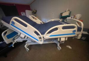 Side view of hospital bed. Bed is hospital-style, not in-home style. Has navy blue mattress, off-white frame with blue accents, and wheeled base. Gently used condition.