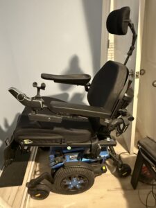 side view of an electric wheelchair with a head rest, arm rests, a joystick controller, and a blue base