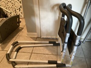 A metal bed assist rail with one u-shaped metal bar designed to lay flat under the mattress, and another u-shaped metal bar sitting vertically with padding on the handles to grab and a fabric storage pouch