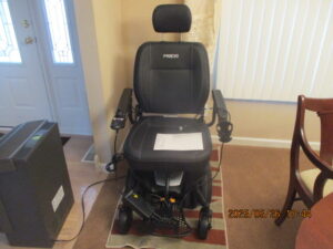 black electric wheelchair with head rest, arms rest and a foot platform. Wheelchair looks like it is plugged in charging.