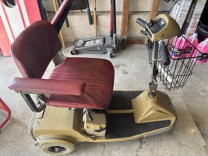a tan three-wheeled mobility scooter with a padded burgundy seat, arm rests, and a wire basket in the front. Scooter has a platform to put feet and handles to hold on to/drive the scooter