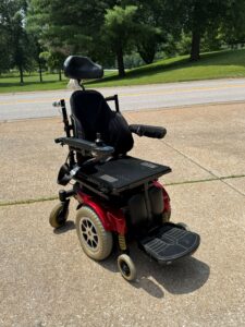 Black power chair with red base. Includes headrest, armrests, footrests, joystick, seatbelt, and attendant handles. Seat cushion is missing. The cover for the backrest is present, but its inner padding may be missing. Fair condition.