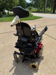 Back view of power chair. A sock is pulled over a piece that extends out from the back of the headrest. A ribbon-like material is wrapped around one of the attendant handles, but it is partly unraveled.