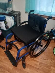 Blue manual wheelchair. Cushion, backrest, attendant handles, footrest, and wheels are black. Backrest appears to have less padding and a different fabric texture from the seat cushion. Gently used condition.