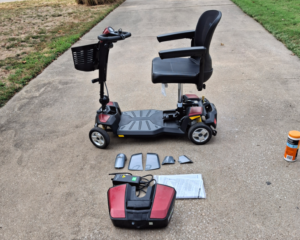 Side view of mobility scooter. Extra items are laid out in front of it, including a manual, charger, GoGo Maxx Li battery, and gray switch plates.