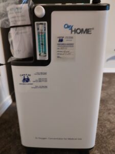 Front view of white oxygen concentrator. Logo says OxyHome. Two informational stickers are on the front, but the picture is too blurry to see them clearly. Item appears to be in good, gently-used condition.