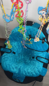 Medium-blue chair with black base. Padded seat cover has a soft, quilted look. A mobile with colorful plastic animals hangs over the chair.