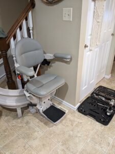 gray seat with arm rests and a foot platform, currently sitting at the bottom of a curved stair rail