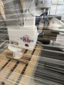Close up of pool lift inside wooden crate. The sides of the crate consist of only a few wooden frames, so the pool lift is visible. Lift is white and gray with an XL Revolution logo. New condition.