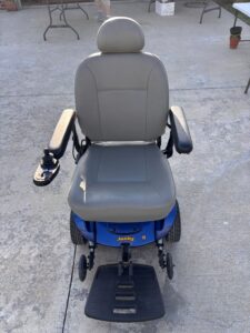 Front view of light grayish-tan power chair with blue base. Chair is padded and has a smooth texture. Includes armrests, joystick, and footrest. Like new condition.