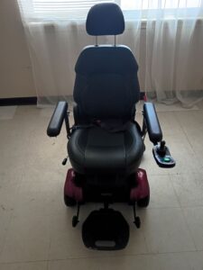 Front view of black power chair with red base. Has control panel, footrest, seatbelt, and cushioned seating. Seating has a smooth texture that may be leather. Gently used condition.