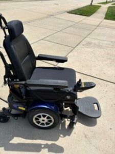 Side view of black power chair with dark blue base. Attendant handles, joystick, footrest, armrests, and headrest are visible. Like new condition. Seat cushion appears to be a different texture than backrest.