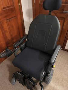 Front view of black power chair. The seat fabric appears slightly different than the back-rest fabric. Seat has a small white smudge and some dust on it, but the rest of the wheelchair is in like-new condition. Head rest, joystick, and footrests are visible.