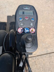 Close-up of power chair’s control panel, located by armrest. Includes joystick, up and down arrows, battery display, volume button, and other buttons.