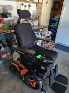 Three-quarters view of power chair. A Jay Fusion label is on the seat cushion. The power chair’s base and inner wheels have orange trim.