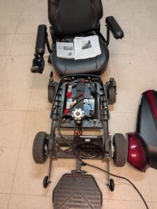 Front view of disassembled power chair. Base is open, revealing inner parts. The word “dynamic” is on one of the parts. The words “Vision CF-P” are visible on top half of base. An owner’s manual and some papers showing the assembly process are on the seat.