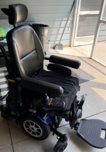 Three-quarter view of power chair, facing right. Attendant handle is visible. Chair appears darker due to shadows.