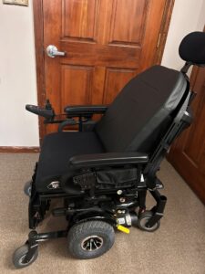 Side view of black power chair, with back rest angled further back. A small side pocket is visible below the arm rest. Chair appears to have six wheels.