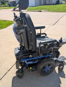 Side view of black power chair with blue base. Chair has cushioned seat, separated footrests, attendant joystick, and control panel with primary joystick. Wheels have some dirt on them.
