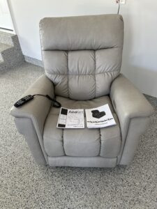 Grayish-beige power recliner. Chair appears to have a smooth fabric texture. An information booklet, an owner’s manual, and a black remote control are on the chair. Like new condition.