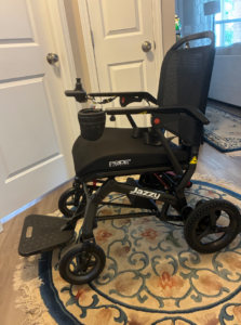 Side view of black, smaller-sized power wheelchair. Includes cup holder, seatbelt, joystick, armrests, and one-piece footboard. Pride Mobility and Jazzy logos are visible. Like new condition.