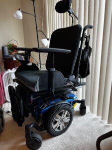 angled view highlighting one side of an electric wheelchair showing the blue base and a large wheel in the middle with a smaller wheel in the front and another smaller wheel in the back. Chair also has handles to push