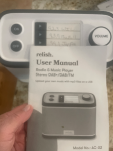 Blurry picture of white radio/music player with gray trim. There is a large on button, off button, and volume knob. Appears to be in like new condition. A hand is holding the user manual over the radio.