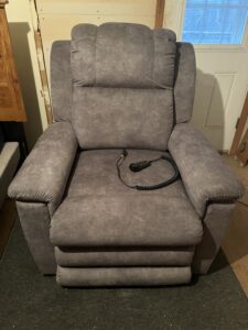 Grayish-beige recliner, shown from front. Like new condition. A black remote control is on the seat. Chair appears to have a soft fabric texture.