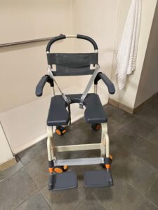 front view of black chair with an opening in the middle of the seat, arm rests, foot rests, and a handle bar on the back to push