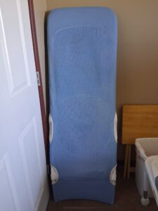 Light blue, rectangular shower chair seat. It is detached from its base and propped up against the wall. Very gently used condition.
