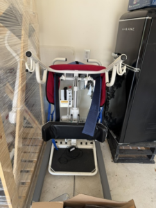 White sit-to-stand device with handles and cushioned back support. A long strip of red and blue material, which may be a strap, is draped over it. Gently used condition.