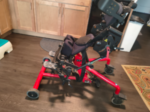 Side view of red sit-to-stand device. Includes wheels, footrests, padded leg supports, padded cushion, padded headrest, and head support. Bantam logo is visible. Gently used condition.