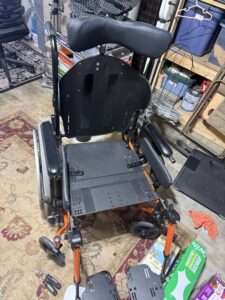 Front view of black wheelchair with footrests. There is no cushioning in the seat area, so the seat’s framework is visible. The parts that connect the seat to the wheels and footrests are orange. Wheelchair appears gently used.