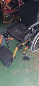 Side of wheelchair, with black cushions added to seat and footrests. Seat cushion has large indentations and looks more worn than the rest of the wheelchair.