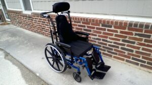 angled view of a manual wheelchair with handles to push, a tall back rest with a head rest, arm rests, and foot holders. Wheelchair has a blue finish at the bottom.