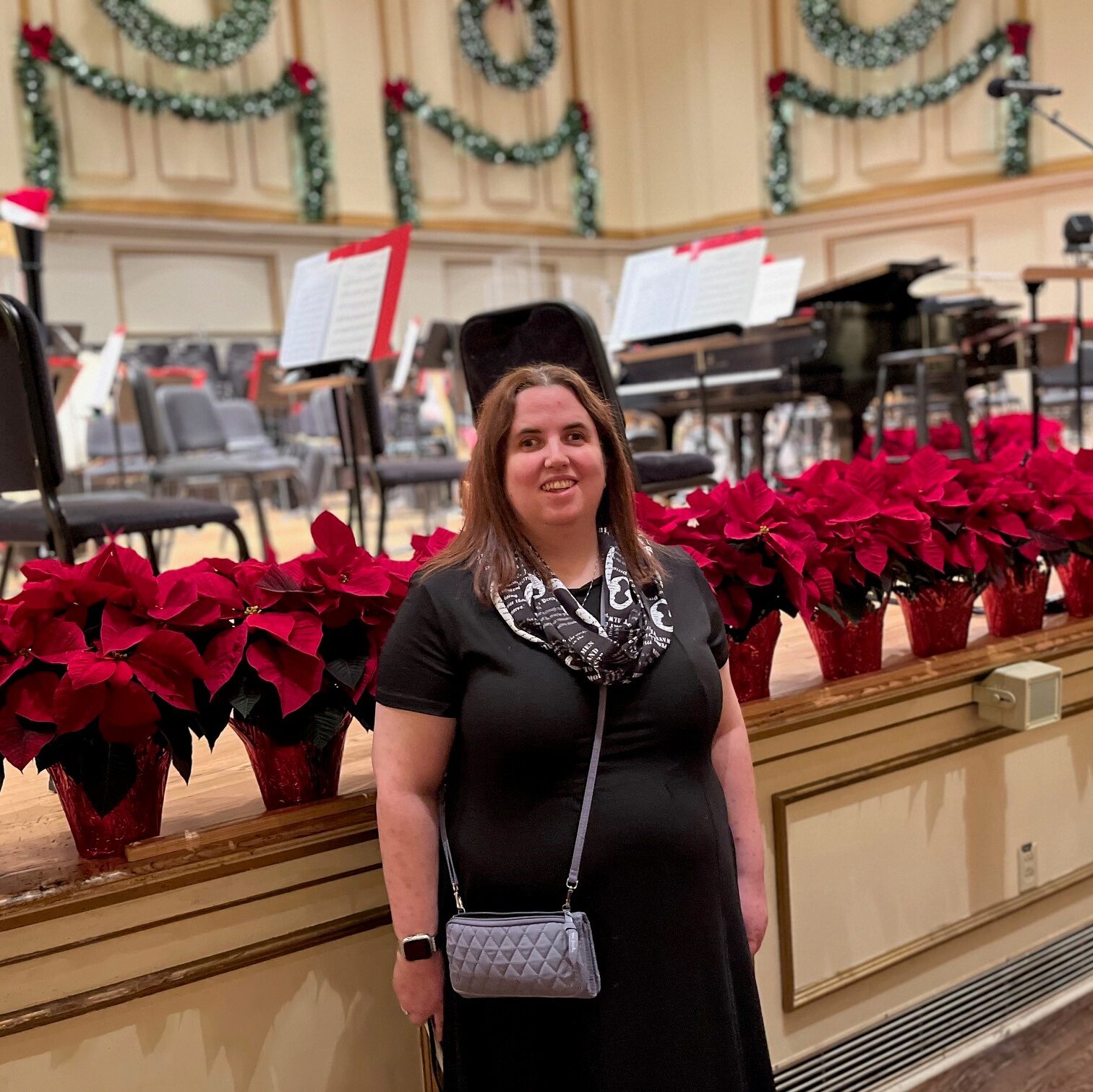 Female dressed in a black dress with gray over the shoulder purse. She is standing in front of a line of red poinsettias.