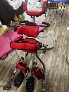 pediatric gait trainer with bright red padding and black straps. Device includes thin frame base with 4 wheels, waist straps, upper body supports, leg straps and foot plates with handles behind to push